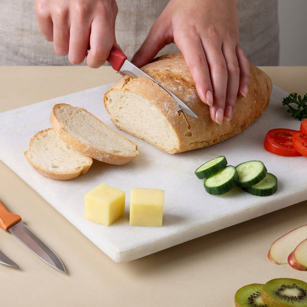 Sure! Here’s the rewritten sentence using your product data: A person slices a loaf of bread on a cutting board with a kitchen prep knife from the Zyliss Classic 3 Piece Peeling & Paring Knife Set. Sliced cucumber, tomato, two cheese cubes, and fruit slices are also neatly arranged on the board next to an assortment of stainless steel Zyliss knives.