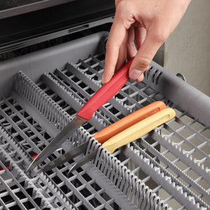 A hand places a red-handled knife from the Zyliss Classic 3 Piece Peeling & Paring Knife Set into the utensil rack of a dishwasher, where two other stainless steel Zyliss knives with orange and yellow handles are already in the rack.