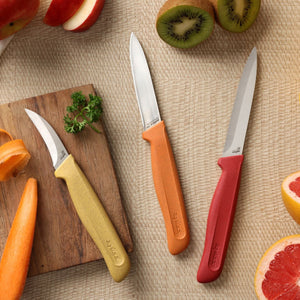 Three stainless steel knives from the Zyliss Classic 3 Piece Peeling & Paring Knife Set, featuring yellow, orange, and red handles, are arranged on a woven surface next to sliced kiwi, grapefruit, and apple. A wooden board with parsley sits nearby. This colorful set is ideal for all your kitchen prep needs.