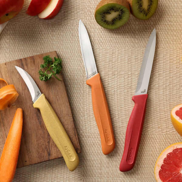 Three stainless steel knives from the Zyliss Classic 3 Piece Peeling & Paring Knife Set, featuring yellow, orange, and red handles, are arranged on a woven surface next to sliced kiwi, grapefruit, and apple. A wooden board with parsley sits nearby. This colorful set is ideal for all your kitchen prep needs.