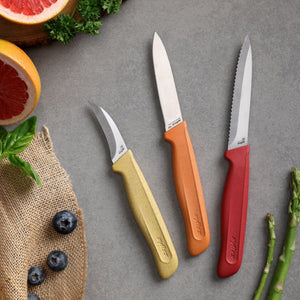 Three kitchen knives from the Zyliss Classic 3 Piece Peeling & Paring Knife Set, featuring yellow, orange, and red handles, are arranged on a gray surface surrounded by blueberries, grapefruit, basil, parsley, asparagus, and a piece of burlap fabric. This stainless steel set brings vibrant color and essential functionality to kitchen prep tasks.