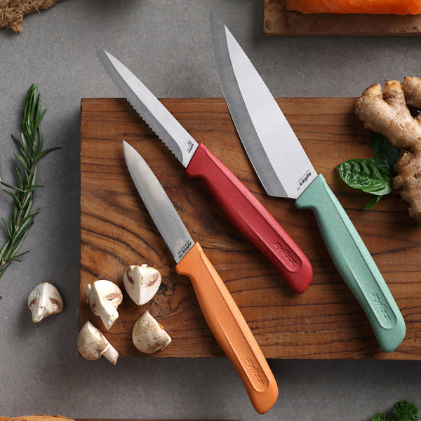 The Zyliss Classic 3-Piece Paring & Utility Knife Set, featuring stainless steel blades and colorful handles, is displayed on a wooden board with mushrooms, ginger, rosemary, and salmon on a separate board.