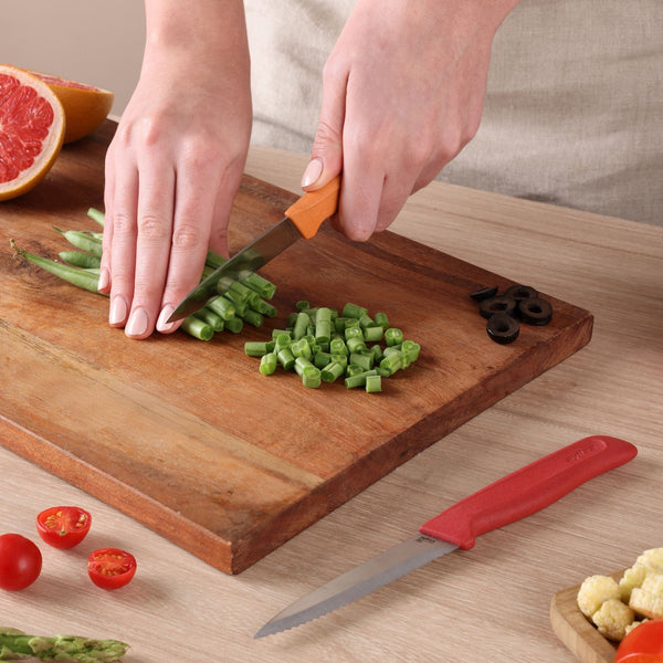 Using the Zyliss Classic 2-Piece Paring Knife Set, a person slices green beans on a wooden cutting board as other ingredients—halved grapefruit, sliced black olives, cherry tomatoes—and a second knife rest on the table.