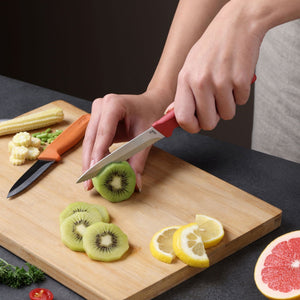 A person slices kiwi on a wooden board using the Zyliss Classic 2-Piece Paring Knife Set with stainless steel blades. Sliced lemon, kiwi, baby corn, green beans, and part of a grapefruit are also on the board.
