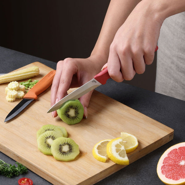A person slices kiwi on a wooden board using the Zyliss Classic 2-Piece Paring Knife Set with stainless steel blades. Sliced lemon, kiwi, baby corn, green beans, and part of a grapefruit are also on the board.