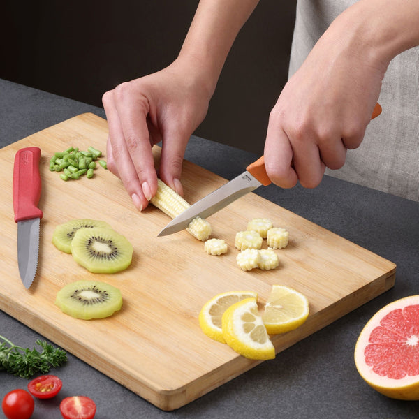 A person slices baby corn on a wooden cutting board with the Zyliss Classic 2-Piece Paring Knife Set, featuring stainless steel blades and ergonomic handles. Chopped green beans, kiwi, lemon, cherry tomatoes, grapefruit, and parsley are nearby.