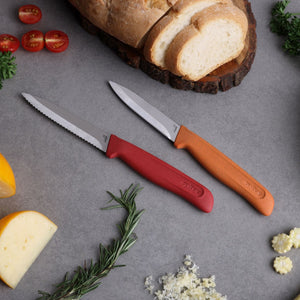 The Zyliss Classic 2-Piece Paring Knife Set with ergonomic handles and stainless steel blades rests on a grey surface, surrounded by sliced bread on a wooden board, cheese, cherry tomatoes, parsley, rosemary, and minced garlic.