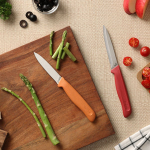 The Zyliss Classic 2-Piece Paring Knife Set, with ergonomic orange and red handles, is displayed on a wooden board surrounded by asparagus, cherry tomatoes, sliced apples, black olives, and a checkered cloth.