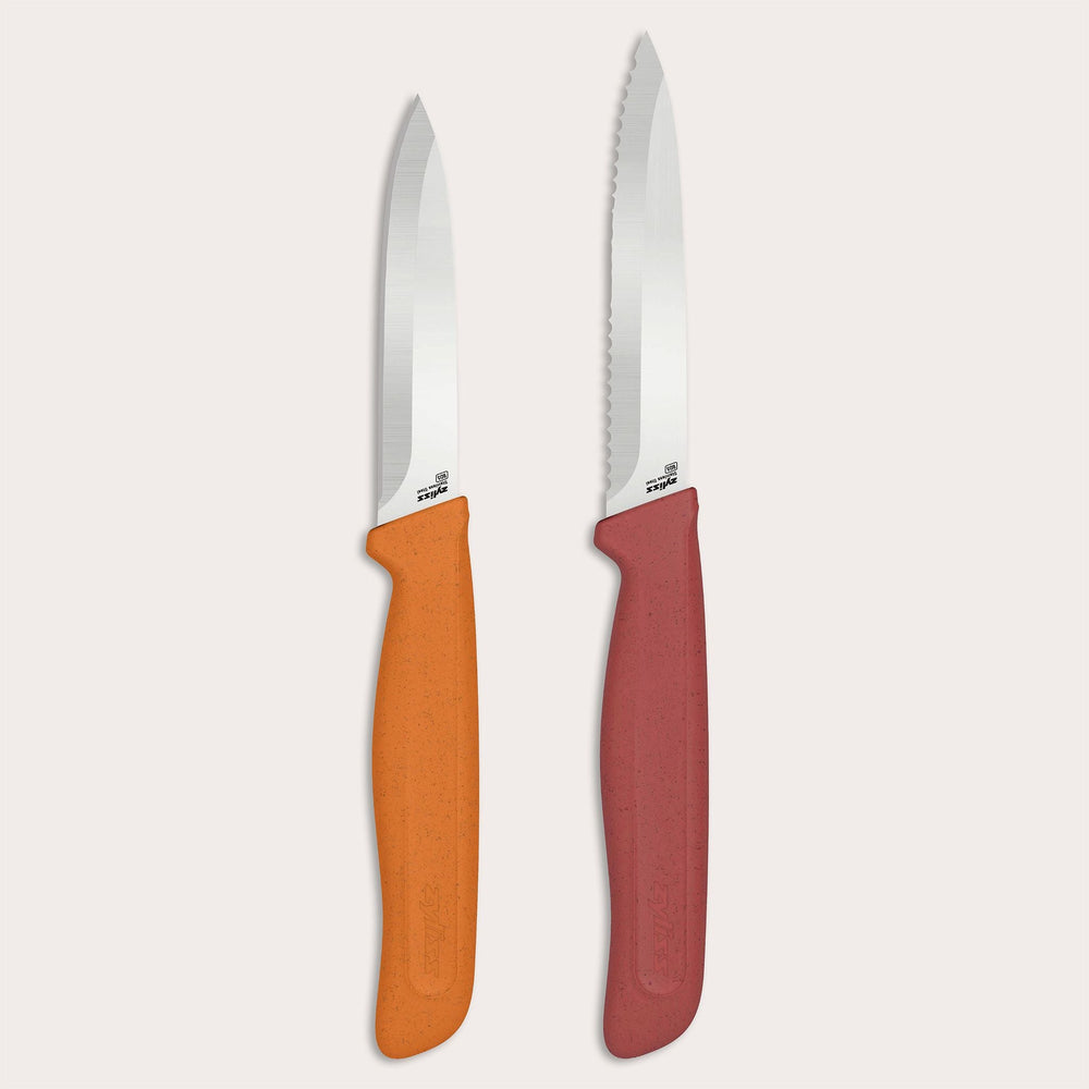 The Zyliss Classic 2-Piece Paring Knife Set features stainless steel blades: one with an orange handle and straight edge, the other with a red handle and serrated blade. Both have textured, ergonomic handles for comfort.