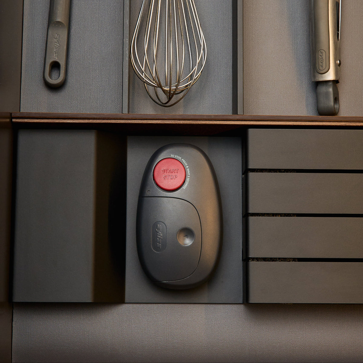 An oval-shaped Zyliss Electronic Can Opener with One-Touch Start/Stop and a red button labeled Start Stop rests in a kitchen drawer organizer alongside other cooking utensils.