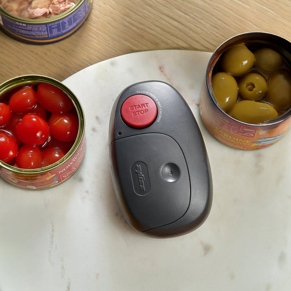 A Zyliss Electronic Can Opener with One-Touch Start/Stop featuring a red button, centered on a marble board between three open cans of food.