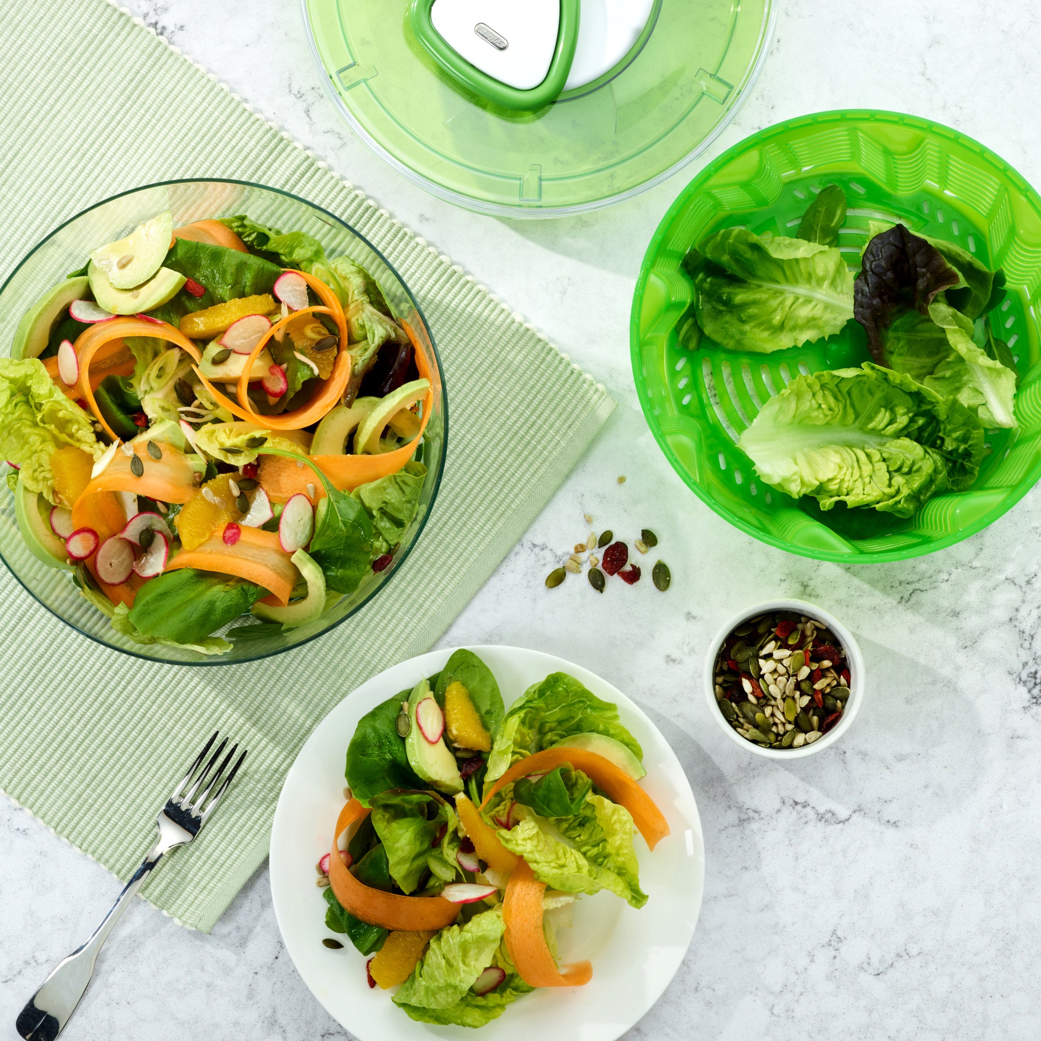 A fresh salad with lettuce, radish, cucumber, and yellow pepper is served in a bowl and on a plate next to the Zyliss Easy Spin 2 Salad Spinner Large with a non-slip base, plus a fork and a small bowl of mixed seeds on a light surface. #White