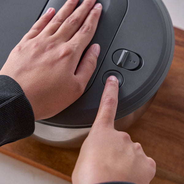 A person presses a button on the lid of the Zyliss Swift Dry Salad Spinner Stainless Steel with one hand while steadying the bowl with the other; the appliance sits on a wooden surface.
