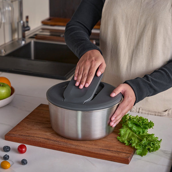 A person in a beige apron places the lid on the Zyliss Swift Dry Salad Spinner Stainless Steel atop a wooden cutting board, surrounded by fresh lettuce, cherry tomatoes, blueberries, and apples in a kitchen.