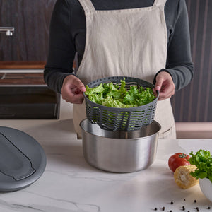 Wearing a beige apron, a person lifts the Zyliss Swift Dry Salad Spinner Stainless Steel basket filled with green lettuce over a bowl on the counter, with vegetables and the one-handed pump lid close by.