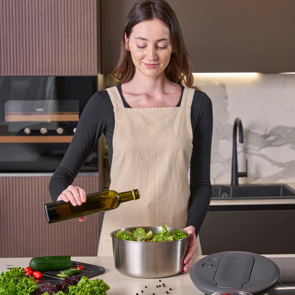 In a modern kitchen, a woman in a beige apron prepares salad beside fresh vegetables, pouring olive oil into the Swift Dry Salad Spinner Stainless Steel by Zyliss.