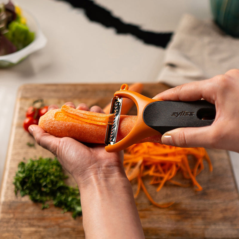 A hand uses an orange vegetable peeler to shred a carrot over a wooden cutting board, with chopped herbs and sliced vegetables visible nearby.