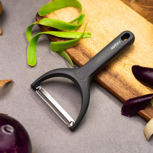 A Zyliss Wide Peeler with an ergonomic handle sits on a gray countertop next to a wooden cutting board, surrounded by green and purple vegetable skins. 