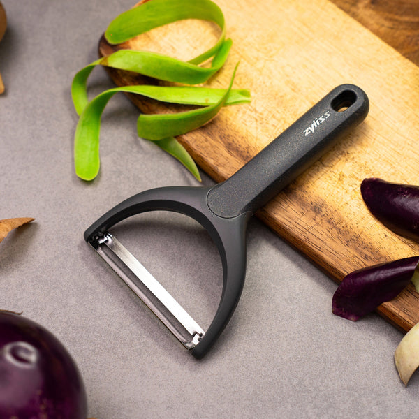 A Zyliss Wide Peeler with an ergonomic handle sits on a gray countertop next to a wooden cutting board, surrounded by green and purple vegetable skins. 