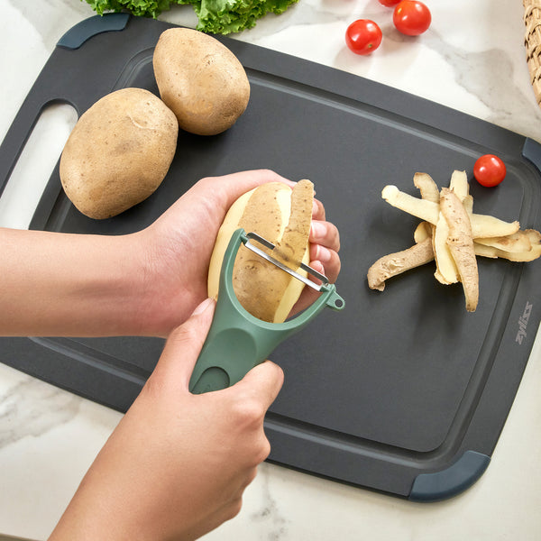 A person uses the Zyliss 3 Piece Y Peeler Set to peel a potato over a black cutting board, with unpeeled potatoes, peels, lettuce, and cherry tomatoes on a light countertop.