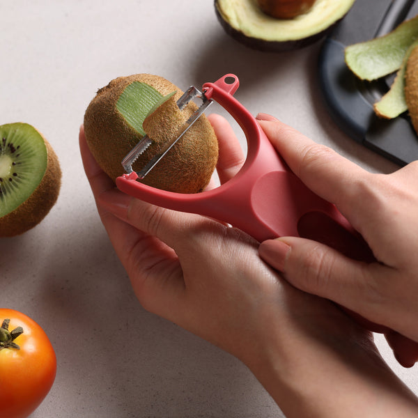 A person uses a red peeler from the Zyliss 3 Piece Y Peeler Set to peel a kiwi, with sliced avocado, tomato, and a black cutting board in the background.