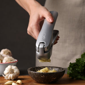 A hand uses a Zyliss garlic press to crush garlic into a bowl on a wooden surface, surrounded by fresh garlic, leafy greens, and components from the Garlic Bread Making Kit. #large