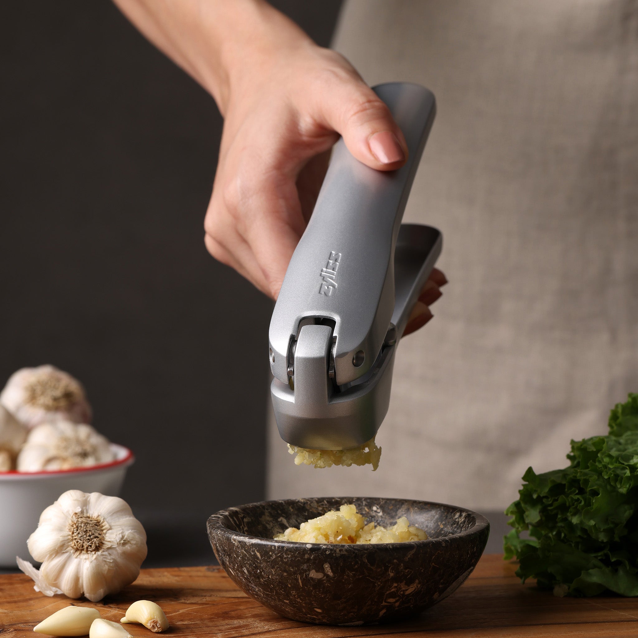 A hand uses a Zyliss garlic press to crush garlic into a bowl on a wooden surface, surrounded by fresh garlic, leafy greens, and components from the Garlic Bread Making Kit. #large