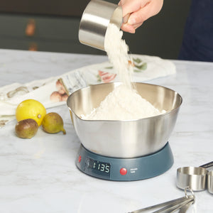 A person pours flour from a metal measuring cup into a bowl on digital kitchen scales with lemons, figs, and other tools nearby—essentials in the Zyliss Banana Bread Baking Essentials Kit.