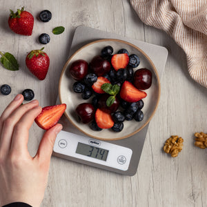 A bowl of mixed berries and cherries sits on a Zyliss Digital Food Scale displaying 374 grams, while walnuts and blueberries are scattered nearby for precise weighing. A hand holds a sliced strawberry above the scale.