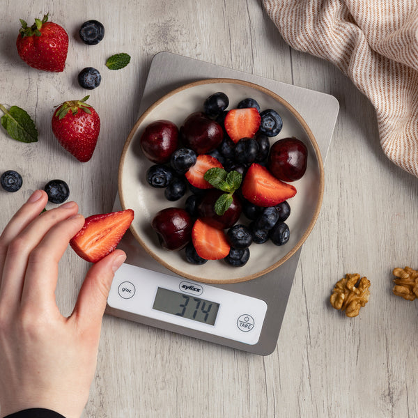 A bowl of mixed berries and cherries sits on a Zyliss Digital Food Scale displaying 374 grams, while walnuts and blueberries are scattered nearby for precise weighing. A hand holds a sliced strawberry above the scale.