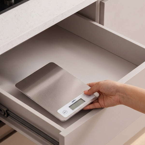 A hand is placing a Zyliss Digital Food Scale with a stainless steel surface into an open kitchen drawer under a white countertop.
