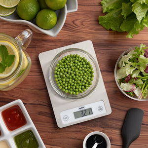 The Zyliss Digital Food Scales with a stainless steel surface accurately display 321 grams of green peas in a bowl, surrounded by fresh salad ingredients and sauces on a wooden table for precise weighing.