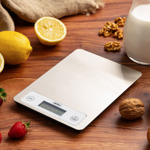 The Zyliss Digital Food Scales with a stainless steel surface rest on a wooden table with strawberries, lemons, walnuts, milk, and a beige cloth. The scale offers precise weighing with its clear display and two easy-to-use buttons.