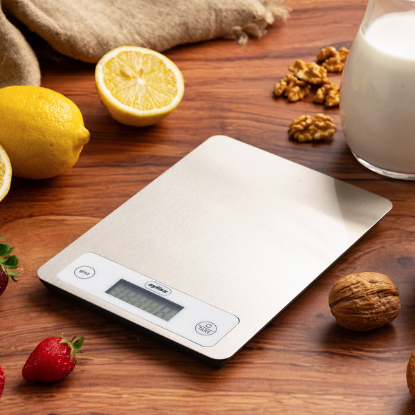 The Zyliss Digital Food Scales with a stainless steel surface rest on a wooden table with strawberries, lemons, walnuts, milk, and a beige cloth. The scale offers precise weighing with its clear display and two easy-to-use buttons.