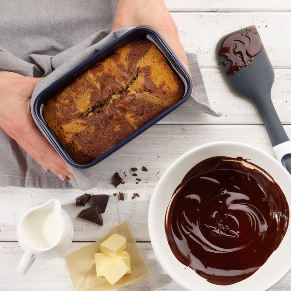 A person holds the Zyliss Banana Bread Baking Essentials Kit's non-stick loaf pan with marble cake, surrounded by a bowl of melted chocolate, a chocolate-covered spatula, milk, butter on parchment, and chocolate chunks on a white surface.