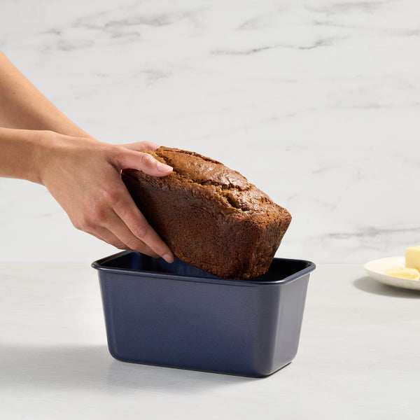 Someone lifts a loaf of banana bread from the Zyliss Banana Bread Baking Essentials Kit’s non-stick pan on a light counter, with a plate of butter in the background.