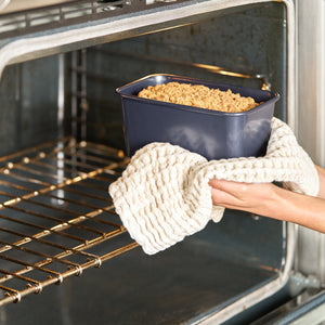 A person uses a textured oven mitt to remove a loaf pan with a baked crumb-topped bread or cake from an open oven. The pan is dark blue and the oven interior is metallic.
