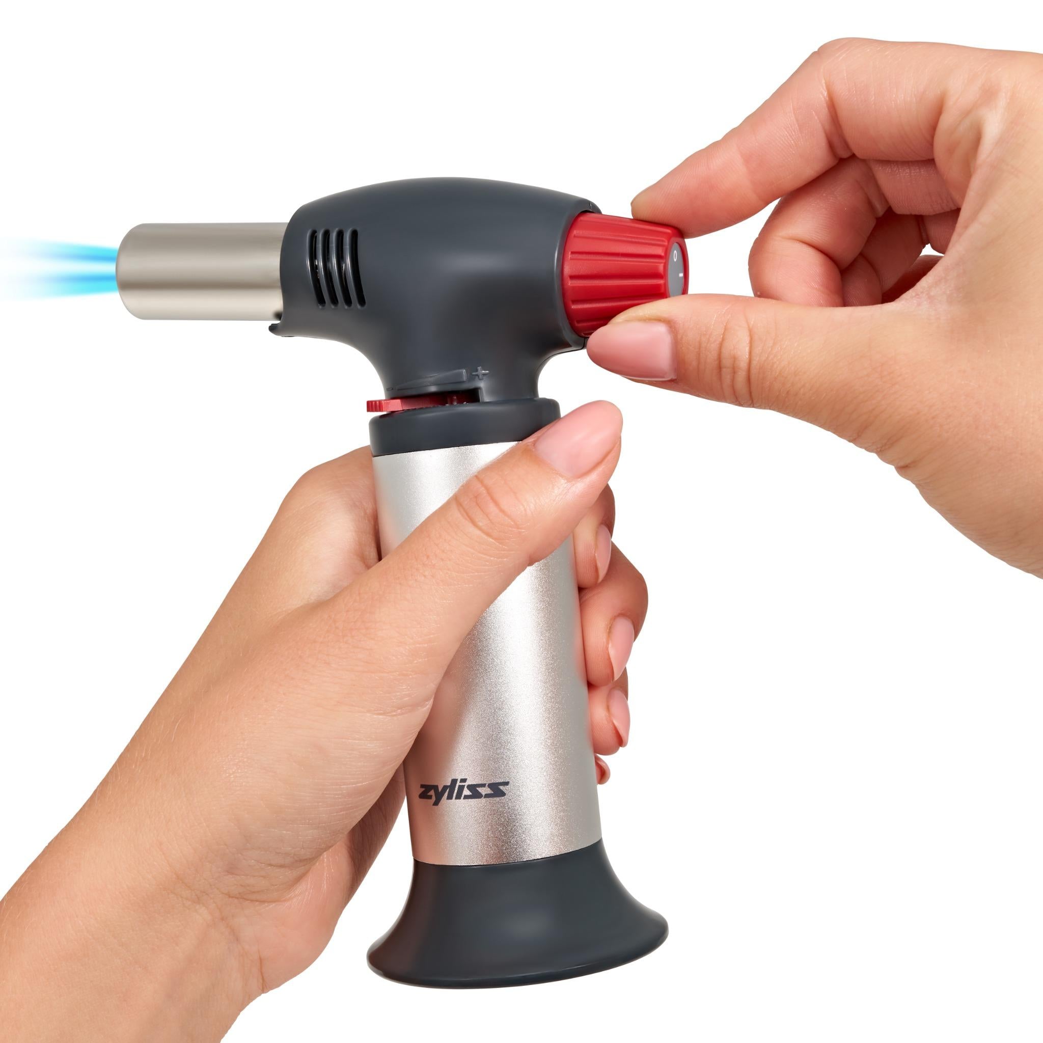 A person holds a silver and black kitchen torch, turning the red ignition knob with one hand while a blue flame comes out of the nozzle. The torch is branded Zyliss.