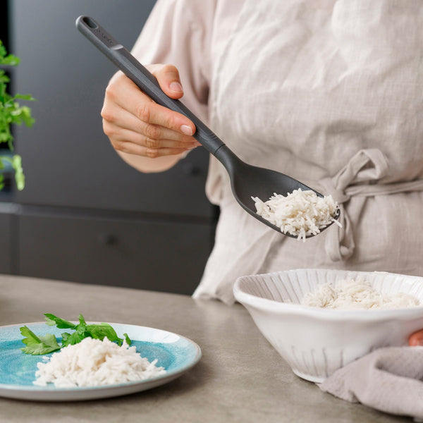 Wearing a light-colored apron, a person uses the Zyliss Spoon to serve cooked white rice from a white bowl onto a blue plate garnished with fresh herbs. #Large
