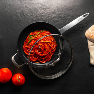 A pan of spaghetti with tomato sauce and basil sits on a black surface. A glass lid partially covers the Zyliss Ultimate Pro Ceramic Saute Pan With Lid, prized for its non-stick coating. Two tomatoes and ciabatta bread wrapped in cloth are nearby. 