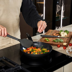 #24cm/9½"
Wearing an apron, a person sprinkles salt over vegetables cooking in a Zyliss Ultimate Ceramic Fry Pan on the stove, holding a spatula, with extra veggies on a cutting board in the background.