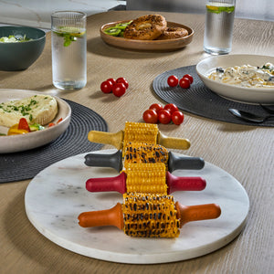 Three grilled corn cobs with Zyliss Corn Holders 4 Piece Set featuring ergonomic stainless steel handles rest atop a marble platter, surrounded by bagels, cheese, cherry tomatoes, salad, and water glasses on a light wooden table.