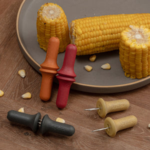 Three cooked corn on the cob pieces rest on a gray plate, each secured with a Zyliss Corn Holders 4 Piece Set featuring ergonomic handles and stainless steel pins, displayed on a wooden surface with scattered kernels.