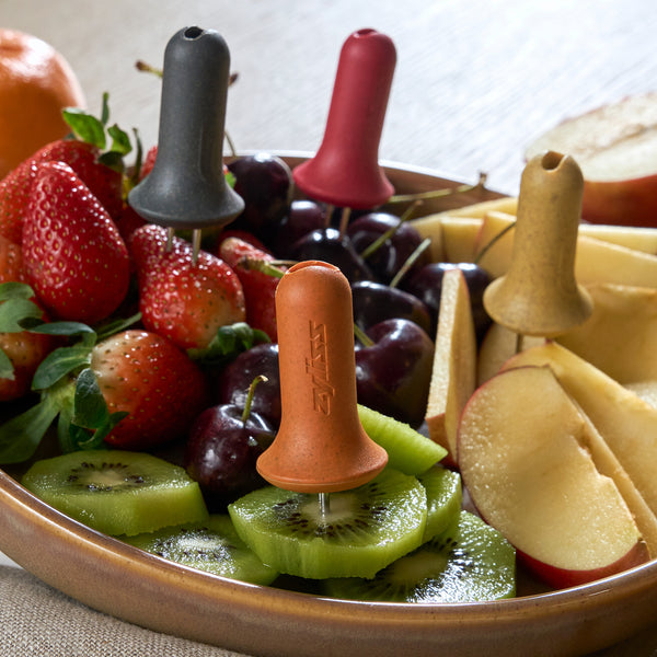 A plate of sliced kiwis, strawberries, cherries, apples, and an orange sits on a table, with Zyliss Corn Holders 4 Piece Set inserted into the fruit pieces as colorful reusable picks.