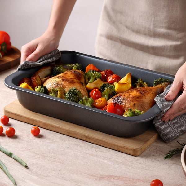 A person uses oven mitts to place a Zyliss Non-Stick Oven Tray with roasted chicken legs, broccoli, cherry tomatoes, and potatoes onto a wooden board on a light countertop.