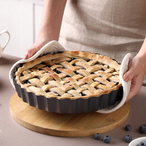 A person holds a freshly baked pie in a Zyliss Non-Stick Tart Pan with removable base, using a white cloth on a wooden board. Blueberries are scattered nearby, and the pie features a woven lattice crust.