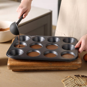 Someone pours chocolate batter into a Zyliss Non-Stick 12 Hole Muffin Pan using a ladle. The pan sits on a wooden cutting board, with waffle biscuits placed nearby on the table.