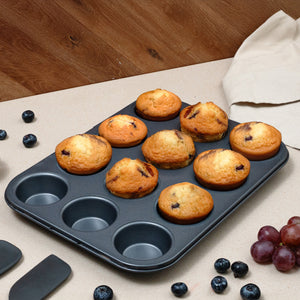 The Zyliss Non-Stick 12 Hole Muffin Pan holds nine baked blueberry muffins, with three empty slots. Blueberries and grapes are scattered on a beige surface, and a cloth napkin is in the background.