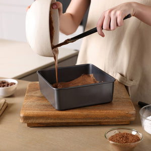 A person pours chocolate cake batter from a bowl into a Zyliss Non-Stick Base Square Pan 20cm / 8" on a wooden board, with cocoa powder and sugar visible on the counter.