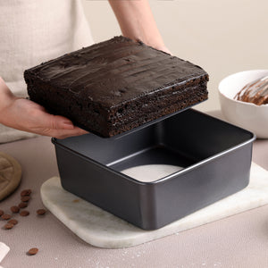 Someone lifts a square chocolate cake layer from the Zyliss Non-Stick Base Square Pan 20cm/8" on a marble board, with a bowl of chocolate batter and scattered chocolate chips nearby.